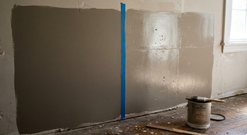 Side-by-side gray paint samples on a wall showing sheen difference, with painter's tape and Benjamin Moore can