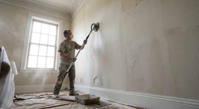 Painter using a pole sander on a skim-coated wall, wearing a mask, with drop cloths on the floor