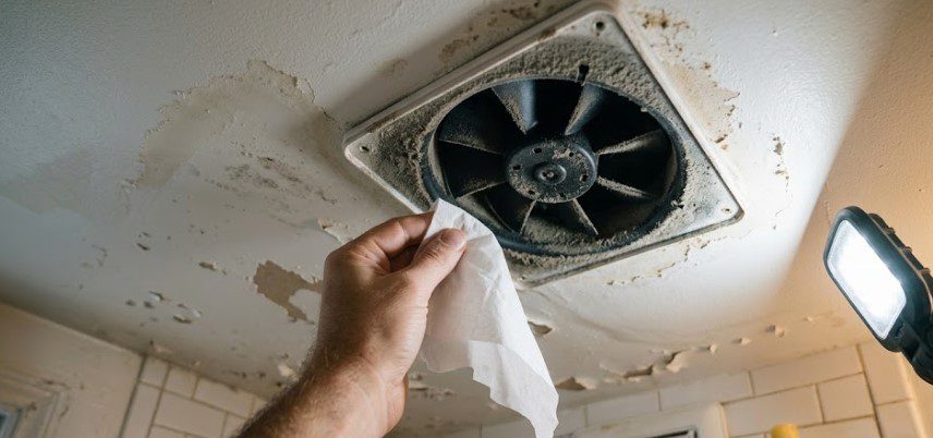 Hand wiping a dusty clogged bathroom exhaust fan with peeling paint around the housing