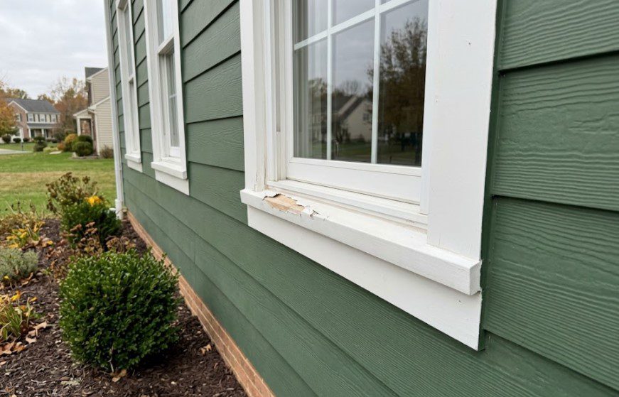 Paint chipped off the corner of a white window sill on a house with green lap siding, exposing bare wood