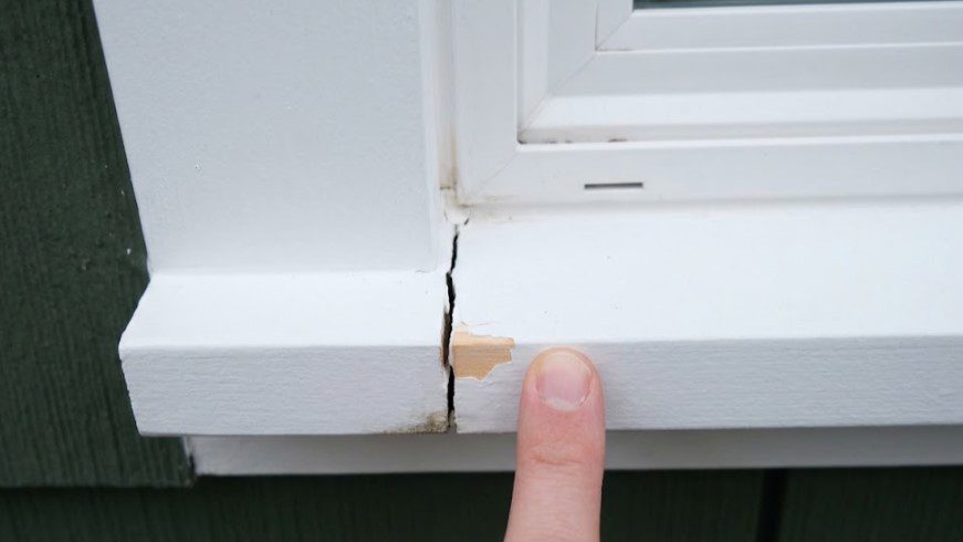 Finger pointing at an open joint between a window sill and its side casing, with chipped paint and dark staining at the bottom of the crack