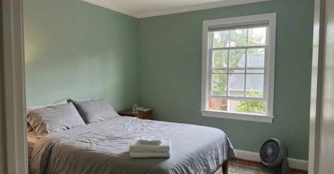 freshly painted green bedroom with the bed made, the lower window sash open and a fan on the floor